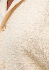 Crème-kleurige gebreide cardigan met een gestructureerd patroon, voorzien van een kraag en twee knopen. Inclusief een subtiele geborduurde logo op de borst.