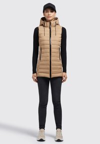 Gilet imbottito beige con cappuccio, chiusura con zip e cuciture verticali. Abbinato con maniche lunghe nere e jeans skinny neri, insieme a scarpe chiare.