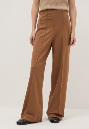 Persoon draagt een broek met hoge taille en wijde pijpen in bruin, gecombineerd met zwarte schoenen en een beige mouwloos gebreid topje, staand tegen een effen achtergrond.