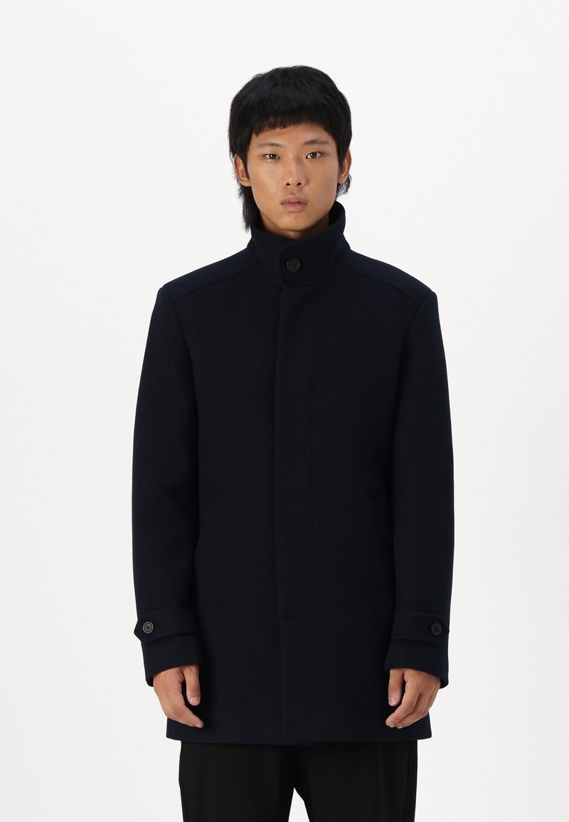 Navy wool coat with high collar, button closure, and side pockets. Features tailored design, long sleeves, and single button cuff details.