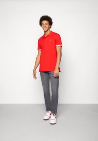Red polo shirt with white striped cuffs, paired with gray jeans and white sneakers featuring red accents. Model stands in a neutral background.