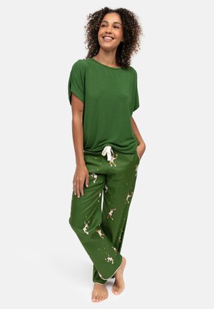 Donna sorridente, indossa una maglietta verde a maniche corte e pantaloni del pigiama verdi con motivo di giocatori di tennis, in piedi a piedi nudi su uno sfondo bianco.