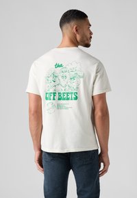 Homme portant un t-shirt blanc avec des personnages de betteraves de dessin animé verts et le texte "The Off Beets" au dos, associé à un jean foncé.