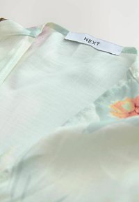 Bluzica s cvjetnim uzorkom napravljena od lagane, prozračne tkanine u svijetlozelenoj boji, s oznakom "NEXT" sašivenom na izrezu vrata.