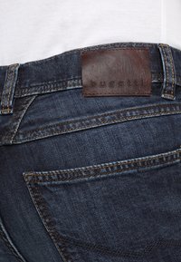 Jeans de mezclilla azul oscuro con una etiqueta de cuero que dice "bugatti" en la parte trasera. Presentan detalles de costura y dos bolsillos traseros.