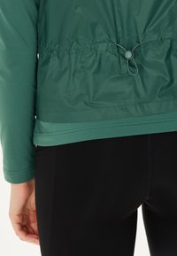 Grön, lätt jacka med dragsko i midjan, lagd över en figurnära svart topp. Funktioner släta texturer och subtila sömmar.