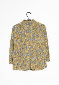 Chemise à manches longues jaune avec un motif abstrait circulaire gris et marron, suspendue sur un cintre noir contre un fond blanc.