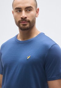 T-shirt bleu à manches courtes en tissu doux avec un col rond et un petit logo oiseau jaune sur le côté gauche de la poitrine.