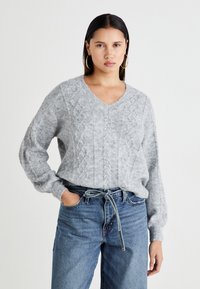 Pull en tricot gris à col en V, manches longues et coupe décontractée, associé à un jean en denim bleu taille haute et une ceinture nouée.