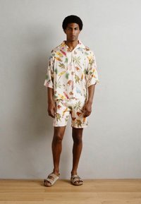 Conjunto de camisa de manga corta con cuello y pantalones cortos en un color beige claro con estampados tropicales coloridos. El modelo lleva sandalias deslizantes marrones.