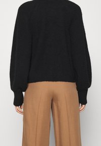 Persona che indossa un maglione nero peloso con maniche a palloncino e pantaloni beige a vita alta e gamba larga, mostrata di spalle.