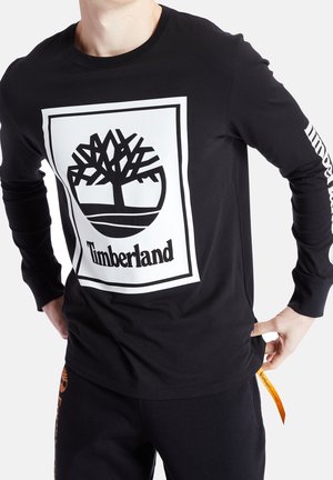 Homme portant un t-shirt Timberland noir à manches longues avec le logo d'un arbre blanc et du texte sur le devant, un marquage sur la manche droite, et un pantalon noir.