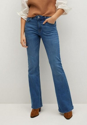 Personne portant un jean flare bleu, des bottines en daim marron et un tricot marron sans manches par-dessus une chemise blanche aux manches retroussées.