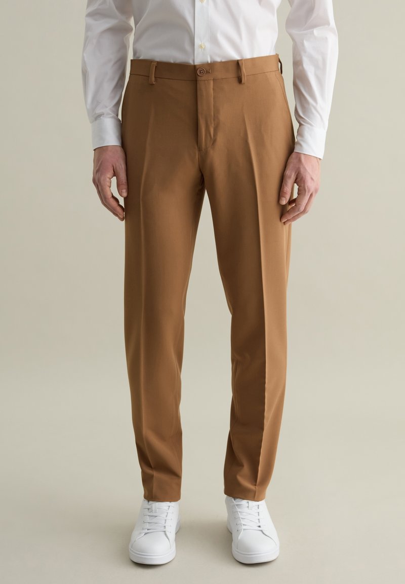 Pantalones tailored marrones con una textura suave, que presentan un cierre de botón y trabillas para el cinturón, combinados con zapatillas blancas.