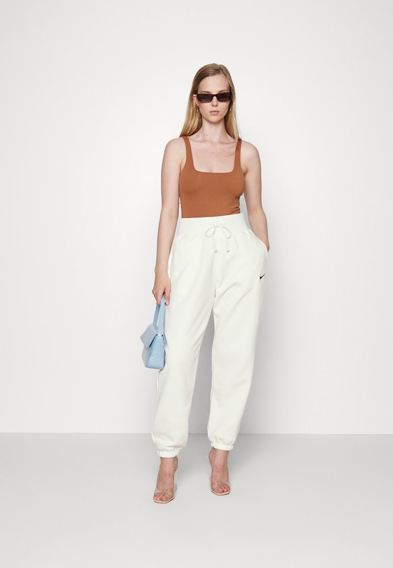 Top de tirantes ajustado marrón, pantalones de chándal blancos con cordón y sandalias de tacón transparente. Un bolso azul añade contraste; diseño minimalista y esquema de colores.