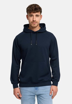 Młody mężczyzna zwrócony przodem, ubrany w granatową bluzę z kapturem z przednią kieszenią i jasnoniebieskie dżinsy na jednolitym białym tle.