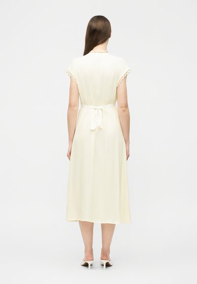 Femme portant une robe midi crème à manches courtes, bordée de dentelle, ceinture nouée à la taille, et des mules blanches à talons, debout devant un fond uni.