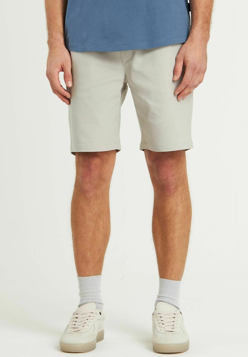Pantaloni scurți gri deschis, fabricați dintr-un material neted, având un design drept și fără buzunare vizibile. Purtați cu adidași albi și șosete gri.