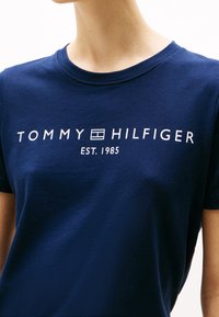 Námořnická modrá bavlněná tričko s bílým potiskem textu "TOMMY HILFIGER EST. 1985" v tučném, moderním písmu. Krátké rukávy a kulatý výstřih.