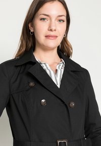 Cappotto trench nero a doppio petto con bottoni marroni, caratterizzato da una vita con cintura e colletto a punte, indossato sopra una camicia a righe.