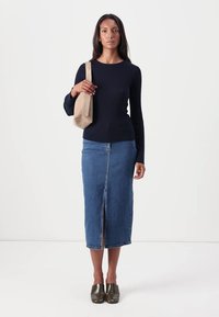 Haut à manches longues côtelé bleu marine, jupe midi en denim avec fente devant, et chaussures métalliques à enfiler ; tenant un sac fourre-tout beige.