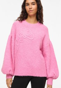 Pull oversize rose en tissu doux et texturé. Présente un motif brodé floral en relief à l'avant ainsi que des poignets et un ourlet côtelés.