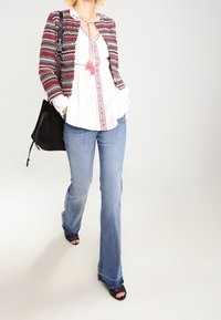 Veste à motifs rouge, blanche et noire sur un chemisier blanc à encolure décorative. Portée avec un jean bleu évasé et des sandales noires.