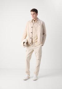 Chaqueta de mezcla de algodón beige con botones negros, combinada con una camisa a juego y pantalones cargo. Unos zapatillas blancas completan el conjunto.