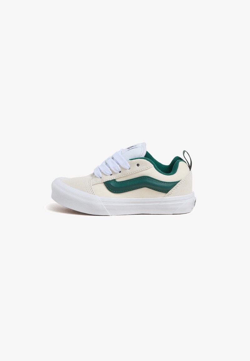 Canvas sneaker in beige with a green accent stripe, white laces, and a rubber sole. Features padded collar and a pull tab on the heel.