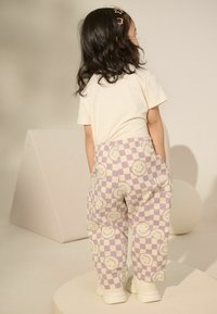 Lila Schachbrett-Hosen mit Mustern von lächelnden Gesichtern, kombiniert mit einem beigen T-Shirt und weißen Turnschuhen. Der Rücken des Kindes ist zur Kamera gewandt.