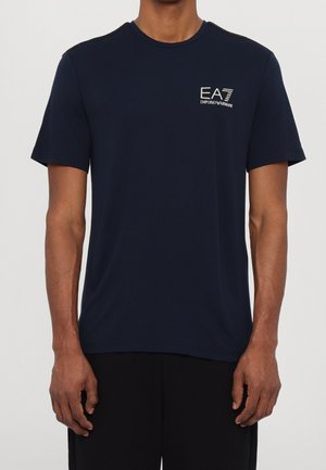 Homme portant un t-shirt bleu marine à manches courtes avec le logo blanc EA7 Emporio Armani sur le côté gauche de la poitrine.