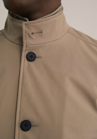 Béžový kabát s vysokým límcem, zdobený černými knoflíky a hladkým, matným povrchem. Obsahuje detaily šití a boční zapínání.