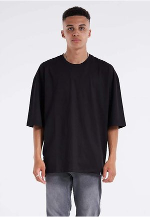Ung mand med kort krøllet hår og fregner iført oversized sort t-shirt og grå jeans, står mod hvid baggrund.
