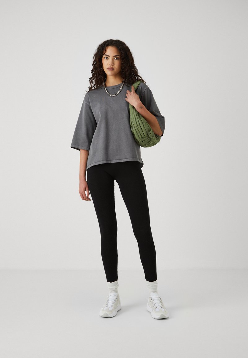 T-shirt gris surdimensionné à manches courtes, associé à des leggings noirs et des baskets blanches. Tenant un sac texturé vert.