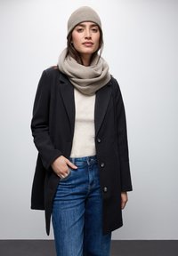Cappotto di lana nero con design doppiopetto, abbinato a una sciarpa e un cappello beige chiaro, indossato sopra un maglione crema e jeans blu.
