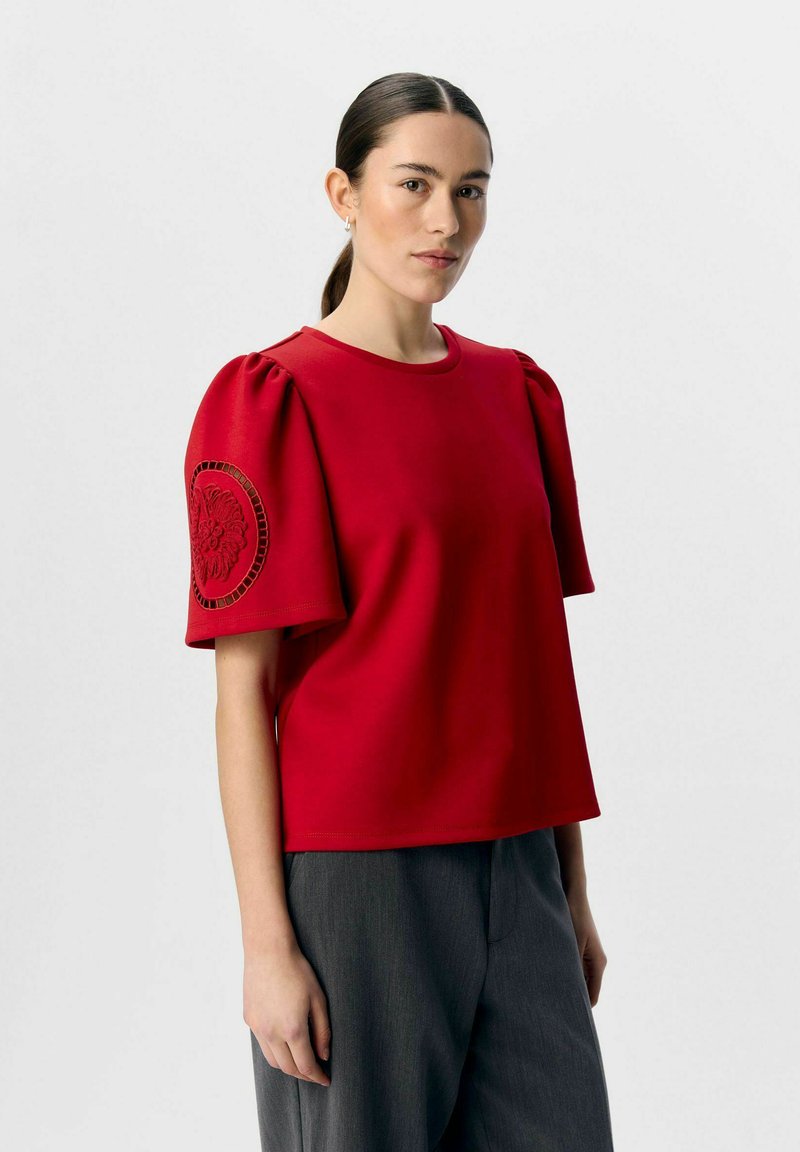 Camiseta roja de manga corta con mangas abullonadas, bordado circular texturizado en la manga izquierda y un cuello redondo. Combinada con pantalones grises.