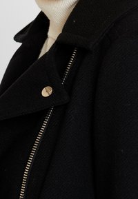 Abrigo de lana negro con cuello con muescas, que presenta un botón dorado y un zipper metálico en la parte frontal. La tela texturizada añade profundidad.