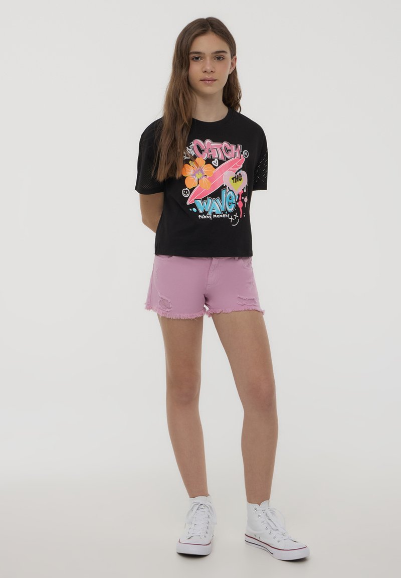 T-shirt nera con design grafico colorato, maniche corte e dettagli in rete, abbinata a pantaloni corti rosa strappati e sneakers bianche.