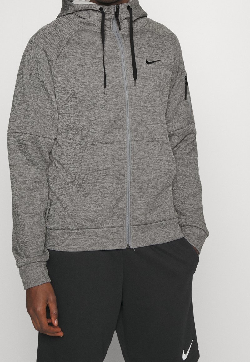 Homme portant un sweat à capuche gris zippé avec des cordons noirs et le logo Nike noir, assorti à un short Nike noir. Les mains détendues le long du corps.