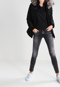 Giacca nera imbottita con cappuccio in pelliccia sintetica, abbinata a un top nero, jeans grigi strappati e sneakers azzurro chiaro.