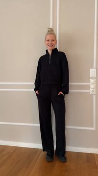 Mujer joven con cabello rubio recogido en un moño, vestida con una camiseta negra con cremallera y pantalones negros, de pie y sonriendo frente a una pared beige.