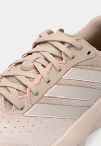 Μπεζ αθλητικό παπούτσι με υφή στο επάνω μέρος, ανοιχτό ροζ λεπτομέρειες, λεία σόλα και τρεις λευκές ρίγες στο πλάι.