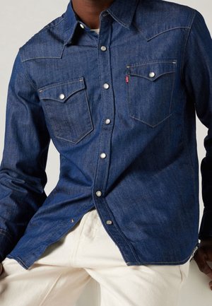 Mand iført en mørkeblå denimskjorte med knapper og to brystlommer samt hvide bukser, mod en ensfarvet baggrund.