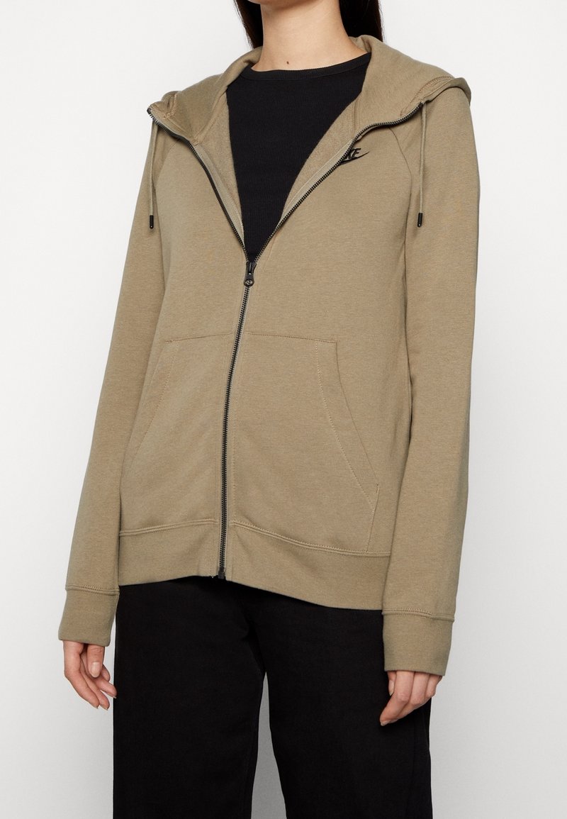 Femme portant un sweat à capuche beige zippé sur un t-shirt noir et un pantalon noir, debout devant un fond clair uni.