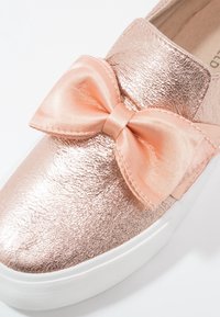 Chaussure slip-on en rose doré métallisé avec un grand nœud en satin pêche sur le dessus, présentant une finition texturée et une semelle en caoutchouc blanche.
