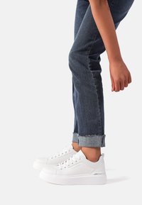 Sneaker bianco con tomaia in pelle liscia, punta rotonda e suola piatta. Abbinato a jeans in denim scuro arrotolati, che mostrano orli sfrangiati.
