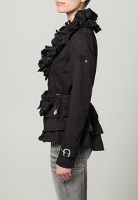 Svart jacka med volanger och lager-på-lager-design, insvängt midja och spänne med grönt inslag. Har texturerat tyg och tydlig krage.