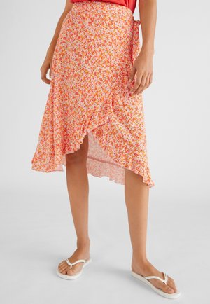 Jupe en fleurs de couleur rose clair avec des fleurs orange et jaunes, ourlet asymétrique, bord froncé, tissu léger, portée avec des tongs blanches.