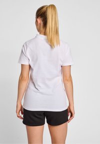 Kvinna med långt blont hästsvans, iklädd en enkel vit t-shirt med korta ärmar och svarta shorts, stående mot en enkel bakgrund.