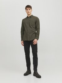 Camisa de botonada verde oscuro con un bolsillo en el pecho, combinada con jeans negros ajustados y botas negras al tobillo, de pie contra un fondo liso.